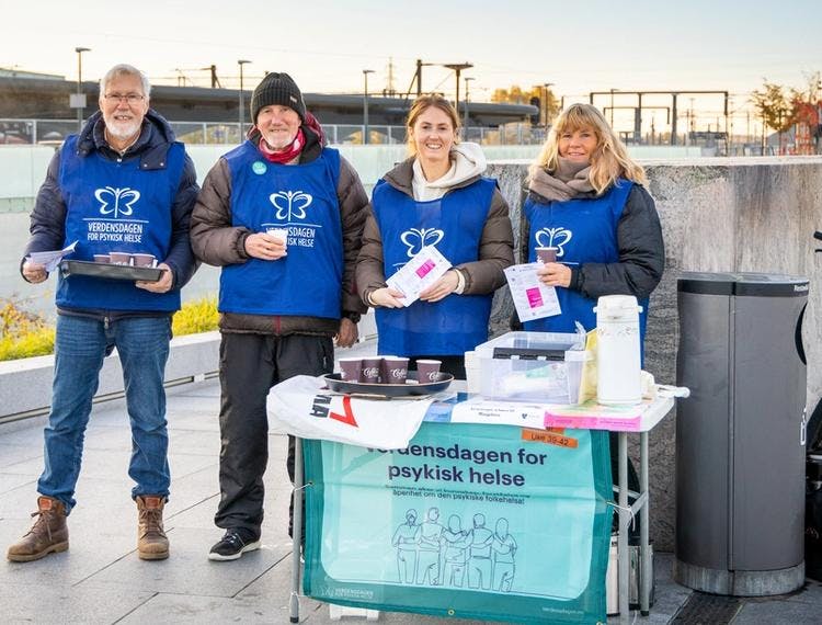 Bilde av fire smilende personer som står på en togstasjon med plakater, bannere, kaffe og vester fra Verdensdagen for psykisk helse